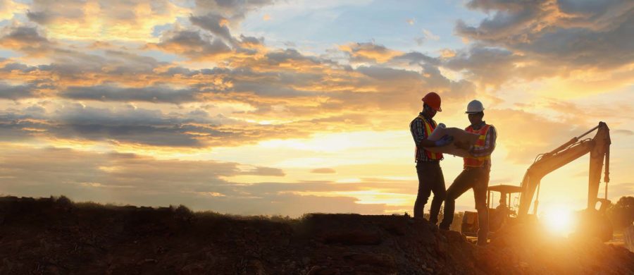 Two construction workers looking over plans, large machinery and the sunrise in the background.