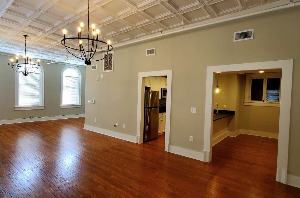 Large room with wooden floors, multiple doors to other rooms. High ceilings, and chandelier lighting. You c an see into the other rooms a little bit.