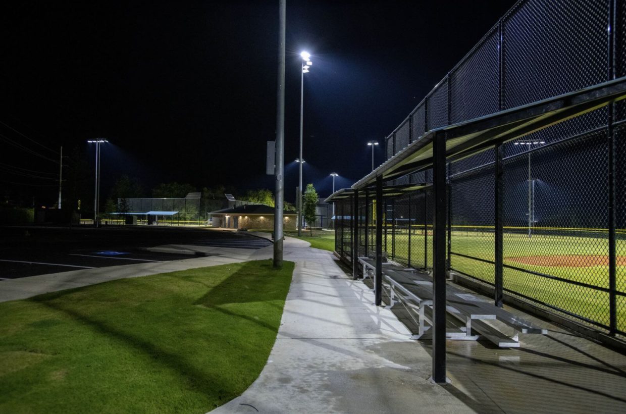 Brick and concrete walkways next to baseball fields, lit up at night, with a small amount of covered stadium seating.
